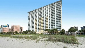 Un hotel alt amb nombrosos pisos, una platja sorrenca al primer pla i altres edificis al fons.