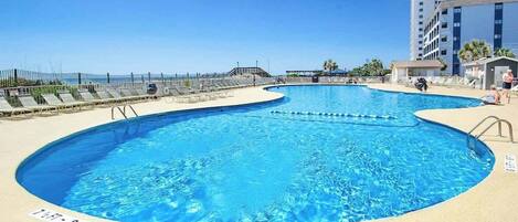 A swimming pool with a curved design, surrounded by a sandy deck and a fence.