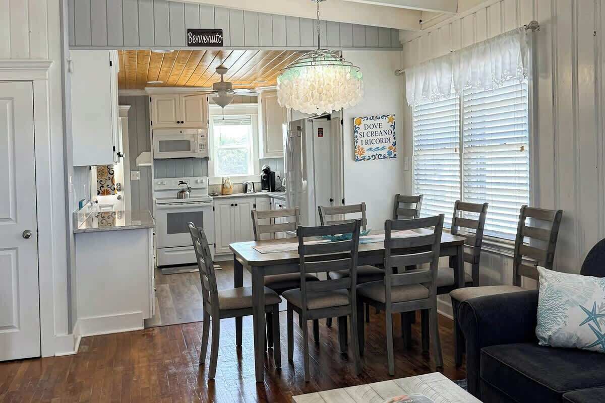 A dining area with a wooden table and chairs, a chandelier, and a kitchen in the background.