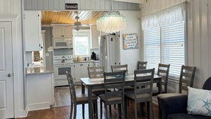 A dining area with a wooden table and chairs, a chandelier, and a kitchen in the background. - Casa del Mare Beach House (North Myrtle Beach)
