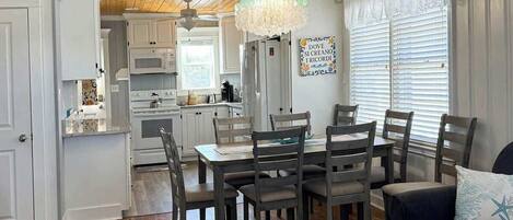 A dining area with a wooden table and chairs, a chandelier, and a kitchen in the background.
