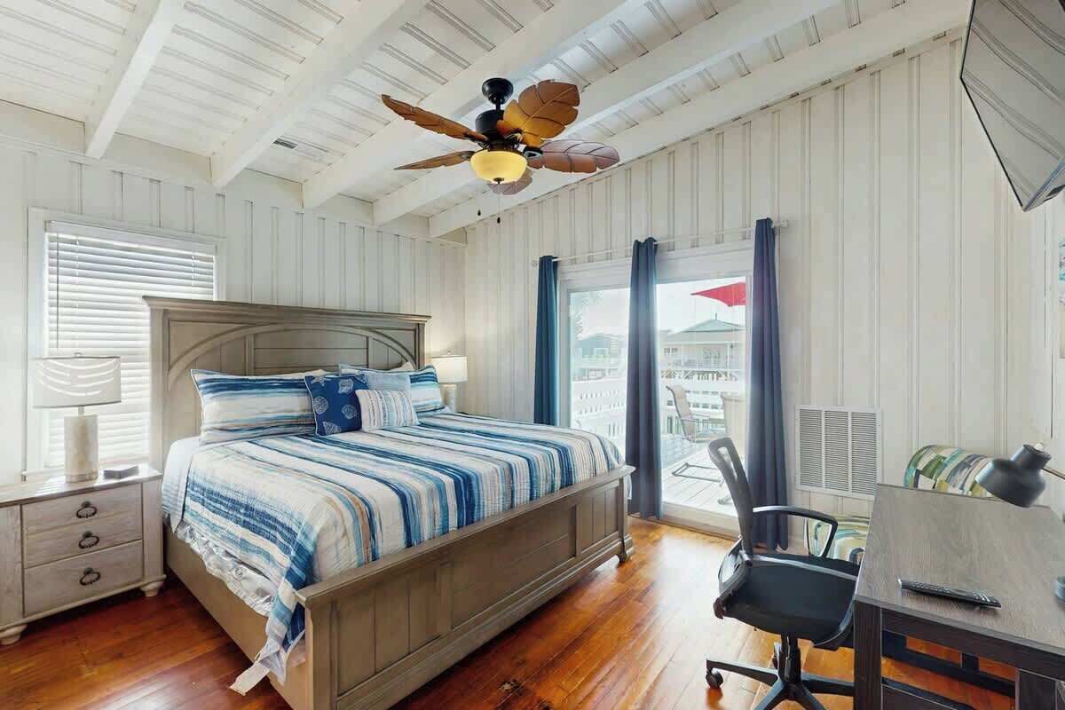 Une chambre à coucher comprenant un lit, un bureau, une chaise, un ventilateur de plafond et une fenêtre avec des rideaux.