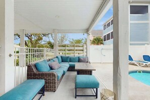 Ein überdachter Außenbereich mit Rattanmöbeln, einer Couch, einem Tisch und einem Pool im Hintergrund.