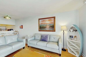 A living room with a sofa, a coffee table, a lamp, and a painting on the wall. - Holiday Towers Unit 704 (Myrtle Beach)