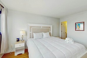 A hotel room with a large bed, a bedside table with a lamp, a wall-mounted painting, and a door leading to another room. - Holiday Towers Unit 704 (Myrtle Beach)