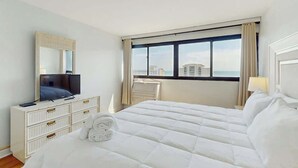 Ein Hotelzimmer mit einem Bett, einem Schminktisch, einem Fernseher, einem Schreibtisch und einem Fenster mit Blick auf Gebäude.