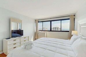 Ein Hotelzimmer mit einem Bett, einem Schminktisch, einem Fernseher, einem Schreibtisch und einem Fenster mit Blick auf Gebäude.