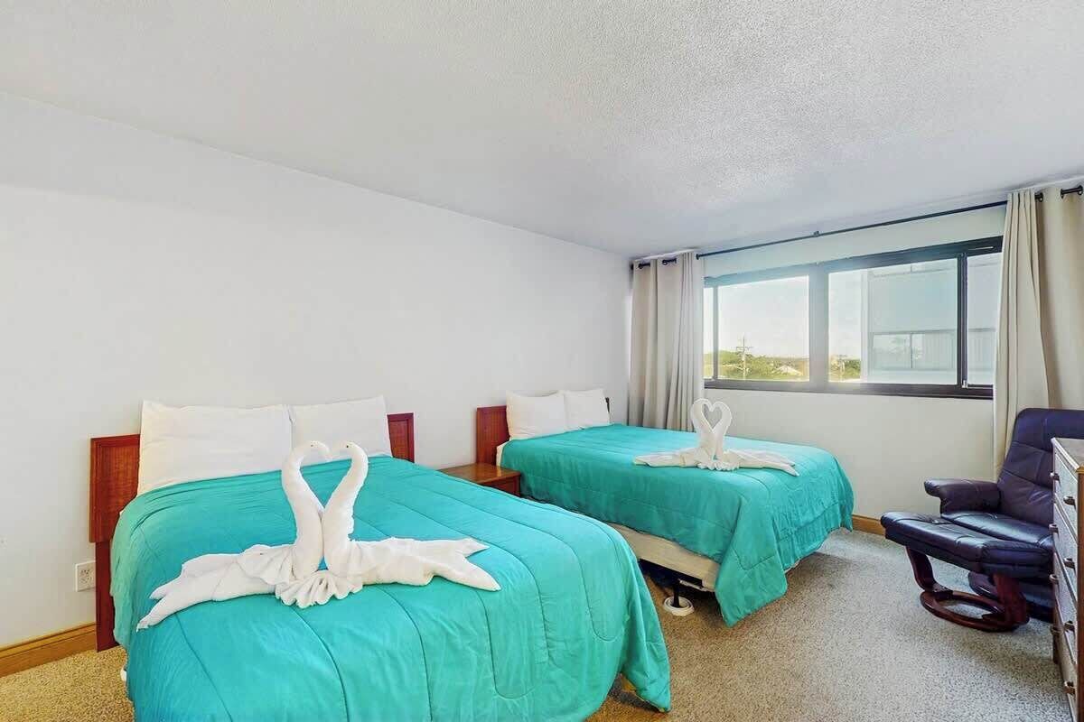 A hotel room with two beds, each with a teal bedspread and white pillows. There is a window with curtains, a chair, and a dresser.