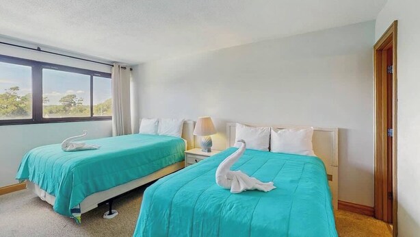 A hotel room with two beds, each with a teal bedspread and a white pillow. There is a window providing a view of trees outside. - Holiday Towers Unit 501 (Myrtle Beach)