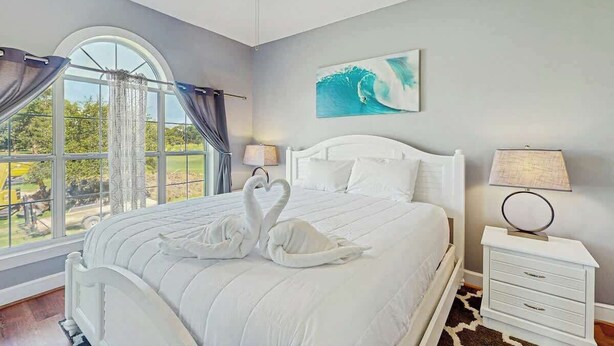 A bedroom with a large bed, a ceiling fan, a window with curtains, a nightstand with a lamp, and a painting on the wall. - Wild Iris Unit 202 Condo (Myrtle Beach)