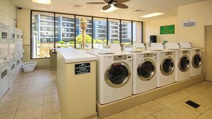 A laundry room with multiple washing machines, a closed sign, and a view of residential buildings through large windows. - Beyond the Blue Retreat (Myrtle Beach)