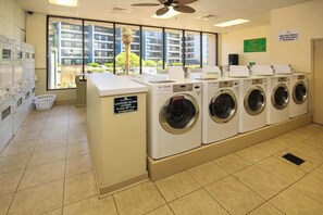 Uma lavanderia com várias máquinas de lavar, um aviso de fechamento e vista para edifícios residenciais através de amplas janelas.