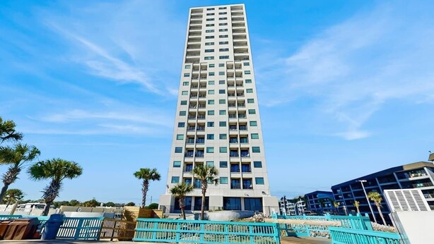 A tall, modern building with a uniform facade and multiple balconies, surrounded by a clear blue sky. - Beyond the Blue Retreat (Myrtle Beach)
