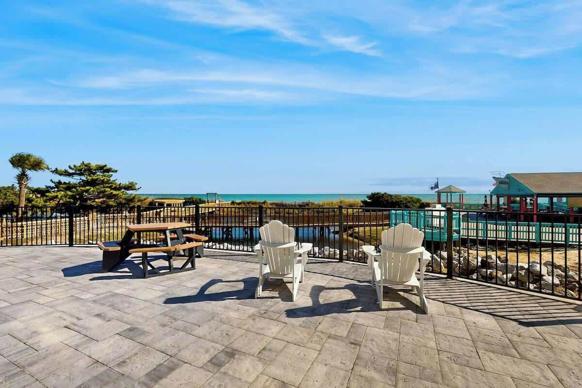 Une terrasse extérieure avec des tables de pique-nique et des fauteuils Adirondack, offrant une vue sur une plage et l’océan.