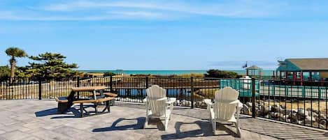 An outdoor patio with picnic tables and Adirondack chairs, overlooking a beach and ocean.