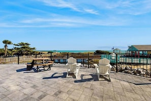 An outdoor patio with picnic tables and Adirondack chairs, overlooking a beach and ocean. - Beyond the Blue Retreat (Myrtle Beach)