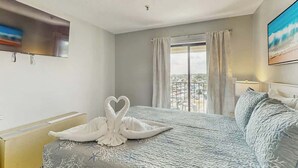A hotel room with a bed, a window with curtains, a flat-screen TV mounted on the wall, and a painting on the wall. - Beyond the Blue Retreat (Myrtle Beach)