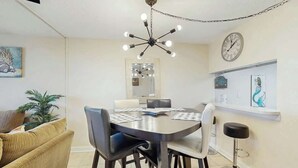 A dining area with a round table, chairs, a chandelier, a clock, and a bar stool. - Holiday Towers Unit 1108 (Myrtle Beach)