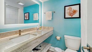 A bathroom with a large mirror, a sink, a toilet, and a framed artwork on the wall. - Grande Cayman Unit 413 (Myrtle Beach)