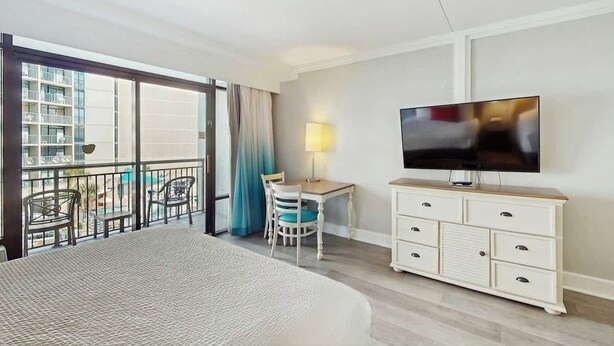 A hotel room with a bed, a TV, a desk with a chair, and a balcony with chairs and a pool view. - Grande Cayman Unit 413 (Myrtle Beach)