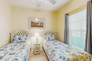 Ein Zimmer mit zwei Betten, einem Deckenventilator, einem Fenster mit Jalousien und einem gerahmten Bild an der Wand.