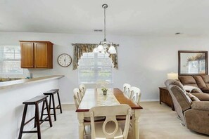 A dining area with a wooden table, white chairs, a chandelier, and a kitchen area with wooden cabinets and a clock. - Willowbend Unit 1511 (North Myrtle Beach)