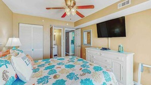 A bedroom with a bed, a ceiling fan, a television, a dresser, and a door leading to another room. - Windy Shores II - 305 (North Myrtle Beach)