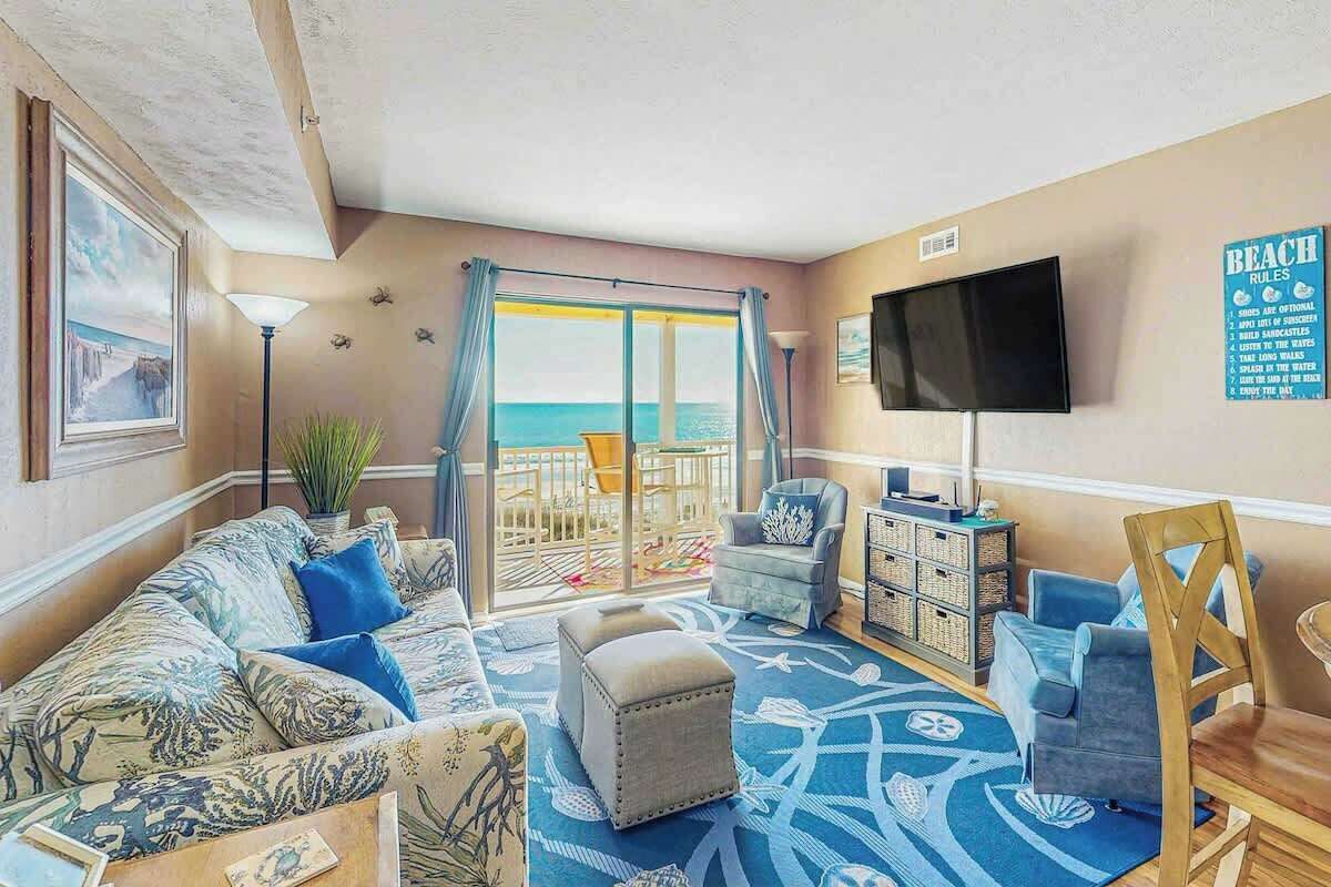 A living room with a view of the beach, a sofa, ottoman, chairs, a TV, and a bookshelf.