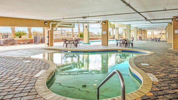 Une piscine intérieure dotée d’un sol carrelé, entourée de chaises longues et de tables, et offrant une vue sur des palmiers et un bâtiment en arrière-plan.