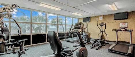 Uma academia com diversos equipamentos de exercício, janelas amplas e uma televisão na parede.