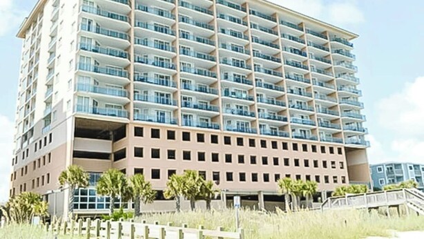 Um hotel com vários andares, varandas, um telhado plano e uma área de estacionamento.