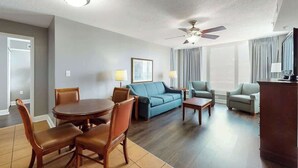Quarto de hotel com área de jantar, sofá, cadeira, ventilador de teto e uma janela grande com cortinas.