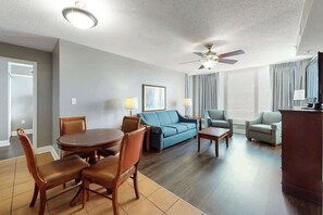 Ein Hotelzimmer mit Essbereich, einer Couch, einem Sessel, einem Deckenventilator und einem großen Fenster mit Vorhängen.