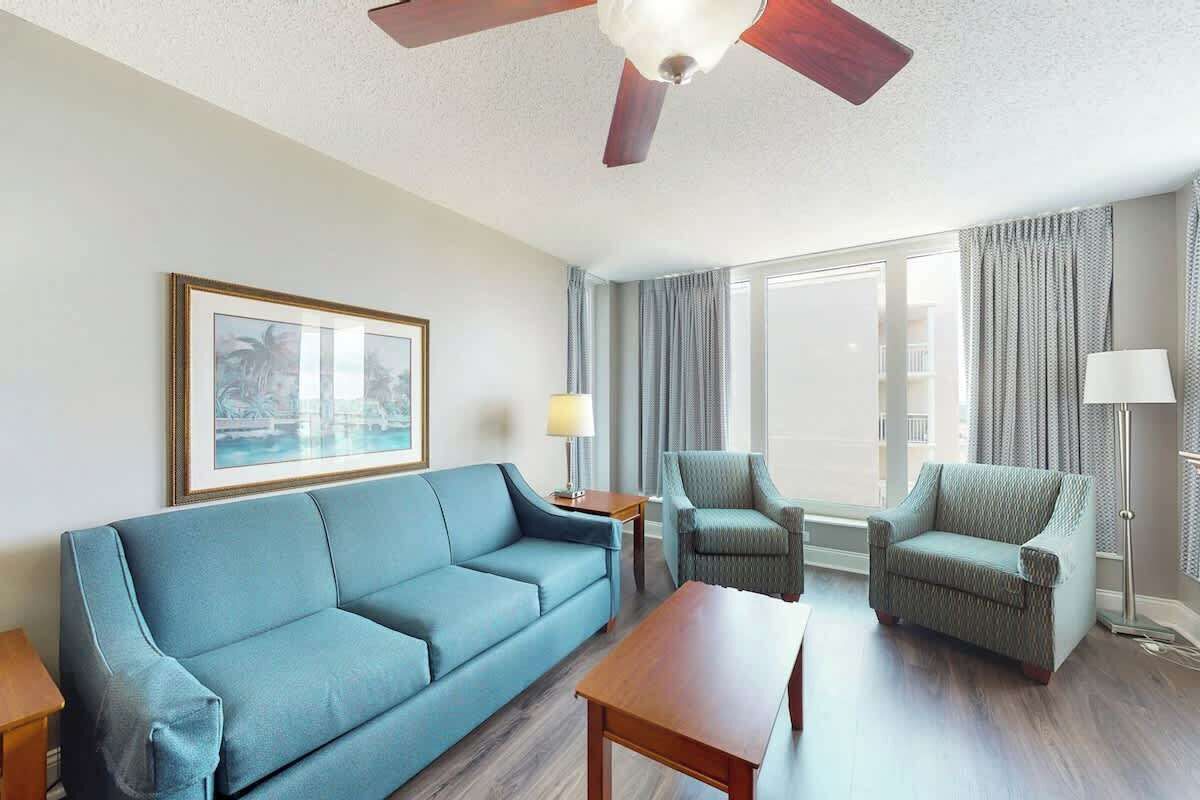 A hotel room with a sofa, two armchairs, a coffee table, a ceiling fan, a framed picture, a lamp, and a large window with curtains.