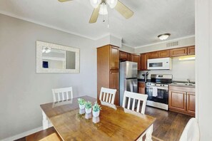 Eine moderne Küche mit Holzschränken, einem Edelstahl-Kühlschrank, einer Mikrowelle und einem Backofen. Ein Esstisch mit Stühlen und ein Deckenventilator sind ebenfalls vorhanden.