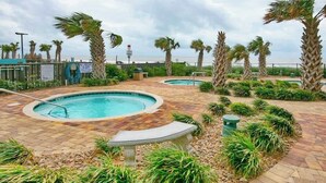Un espace piscine doté de deux bains à remous, entouré de palmiers et de plantes, et offrant une vue sur l’océan.