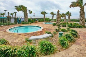 A pool area with two hot tubs, surrounded by palm trees and plants, with a view of the ocean. - Palace Resort Unit 706 (Myrtle Beach)