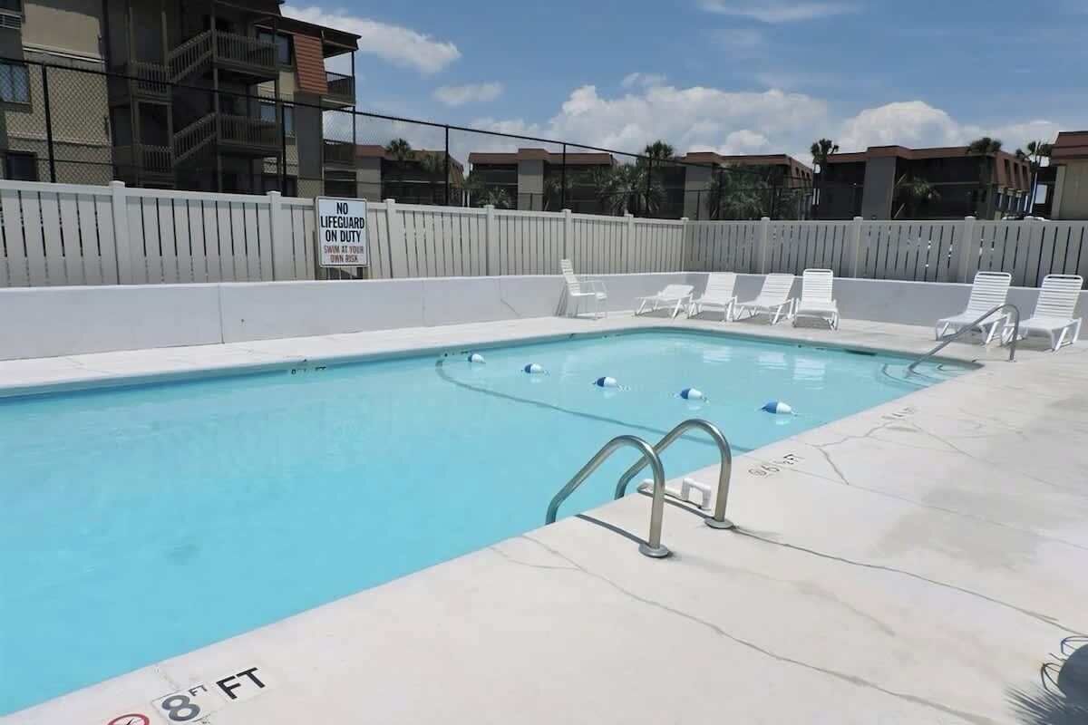 Una piscina con un cartel de no haber socorrista, rodeada de sillas reclinables blancas y una cerca blanca.