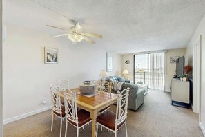 A dining area with a table and chairs, a ceiling fan, a sofa, a TV, and a window with a view. - Holiday Towers Unit 607 (Myrtle Beach)