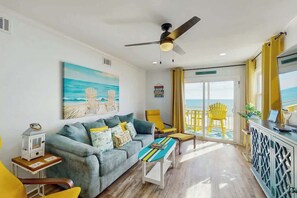 Ein Zimmer mit einer Couch, einem kleinen Tisch, einem Deckenventilator und Blick auf den Strand durch eine Glastür.