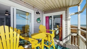 A covered outdoor area with yellow chairs and a table, a glass door leading to a beach view, and a small porch with a railing. - Windsong Unit 303 Condo (North Myrtle Beach)