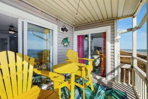 A covered outdoor area with yellow chairs and a table, a glass door leading to a beach view, and a small porch with a railing. - Windsong Unit 303 Condo (North Myrtle Beach)