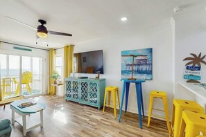 Uma sala de estar luminosa, com televisão, poltronas amarelas, uma mesa azul e um quadro de um cais.