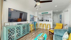 A modern living room with a large flat-screen TV, a teal cabinet, yellow bar stools, a blue sofa, and a ceiling fan. - Windsong Unit 303 Condo (North Myrtle Beach)