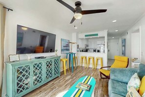 A modern living room with a large flat-screen TV, a teal cabinet, yellow bar stools, a blue sofa, and a ceiling fan. - Windsong Unit 303 Condo (North Myrtle Beach)