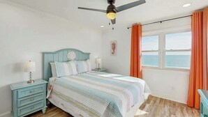 A bedroom with a bed, nightstands, a ceiling fan, a window with curtains, and a view of the sea. - Windsong Unit 303 Condo (North Myrtle Beach)