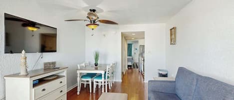 A living room with a grey sofa, a dining table with chairs, a ceiling fan, and a TV mounted on the wall.
