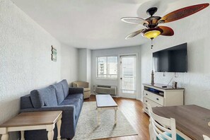 A compact living space with a sofa, a small table, a TV mounted on the wall, and a ceiling fan. - Bluewater Resort 1006 (Myrtle Beach)