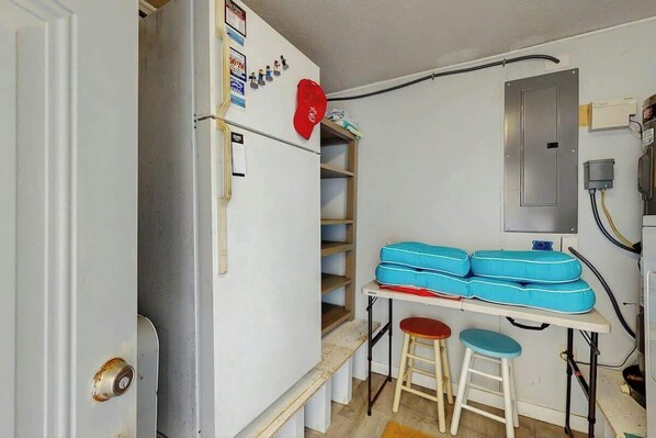 Ein kleiner Raum mit weißer Tür, einem Tisch mit blauen Kissen, zwei Hocker und einem Regal mit verschiedenen Gegenständen.
