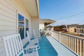 A covered porch with white chairs and a blue floor, overlooking a street with houses. - Ward's Landing Retreat (North Myrtle Beach)